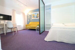 Quarto de hotel moderno com cama, escrivaninha com computador, sofá amarelo e um quadro na parede.