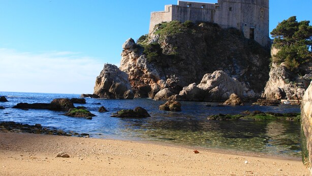 Un castillo costero en un promontorio rocoso con agua azul cristalina y una playa de guijarros al frente.