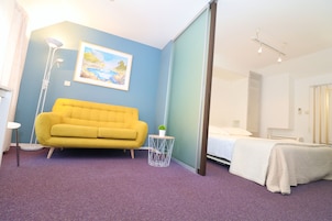 A room with a yellow sofa, a bed with a white blanket, a small round table with a plant, a floor lamp, and a painting on the wall.