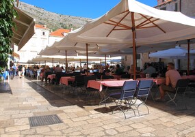An outdoor café with tables and chairs under white umbrellas, set against a mountainous backdrop.