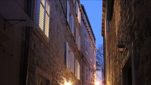 Una estrecha calle adoquinada flanqueada por edificios de piedra con numerosas ventanas y puertas, iluminada por una cálida luz.