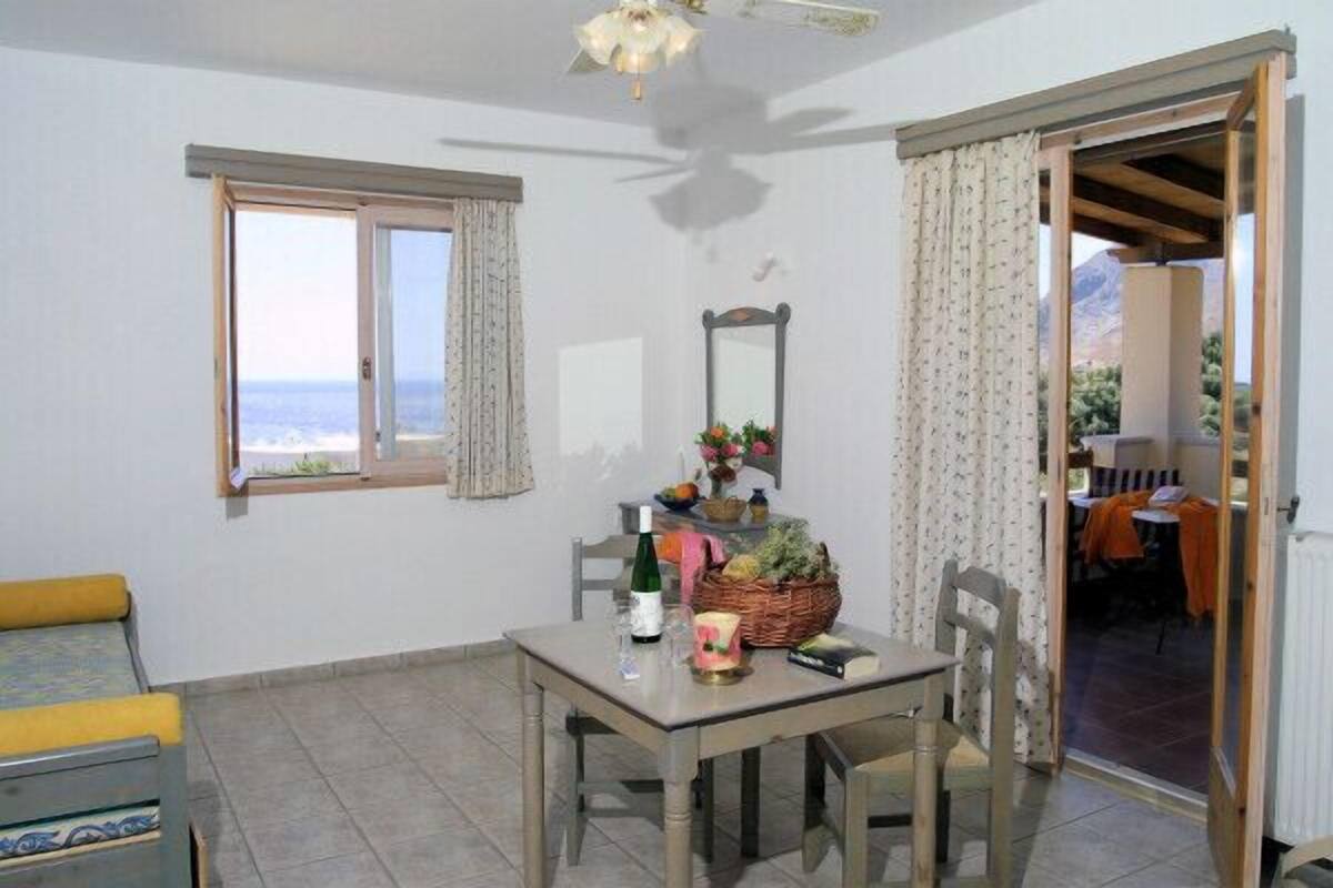 A room with a dining table, chairs, a small sofa, a window with curtains, a mirror, and a view of the sea.