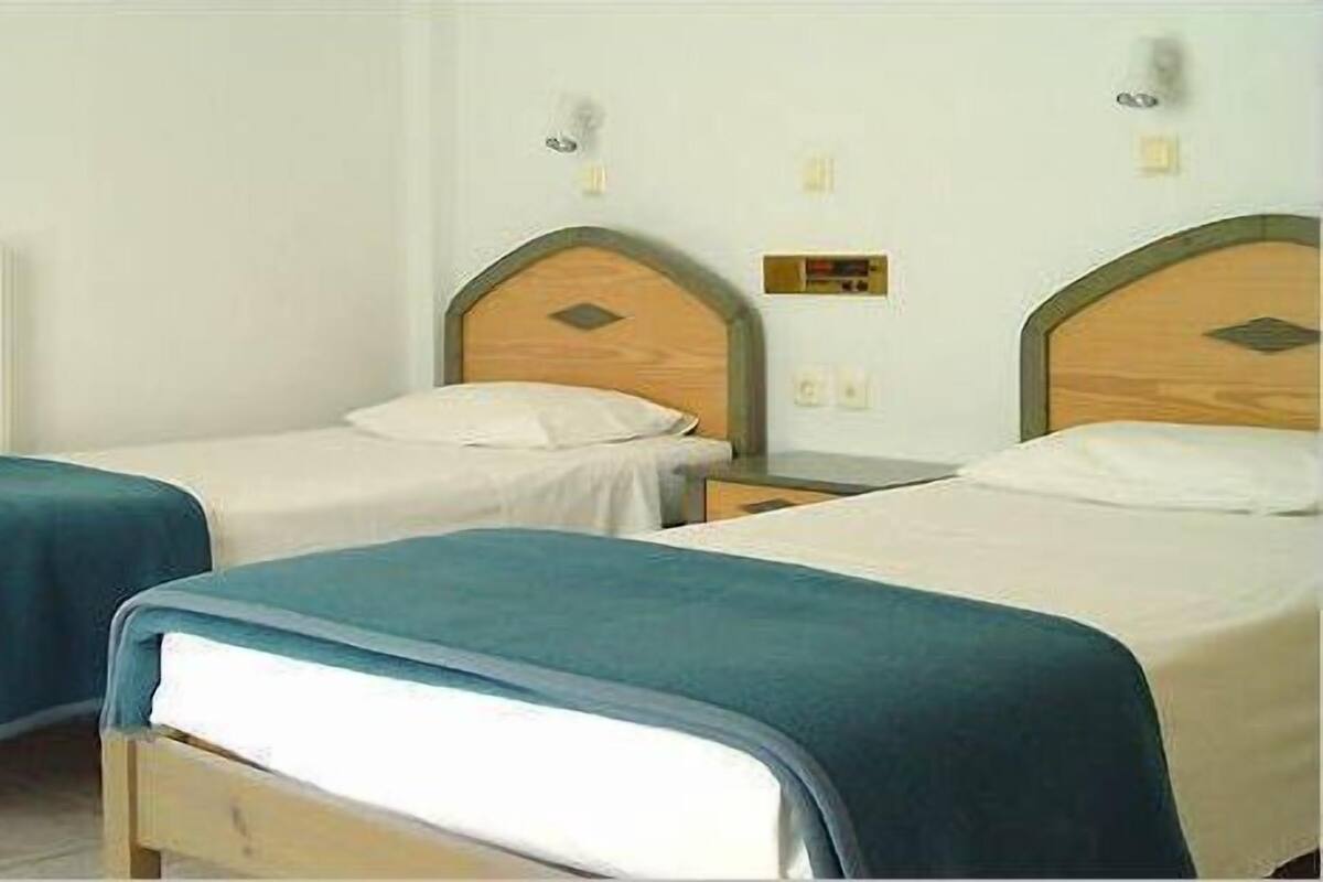 A room with two single beds, each with a wooden headboard and a white mattress. There are bedside tables with lamps and a small shelf. The walls are plain white, and there is a radiator on the left side.