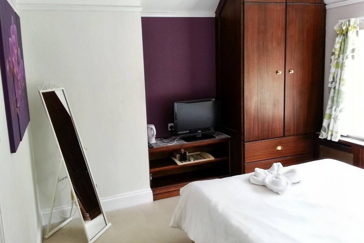 A hotel room with a bed, a wooden wardrobe, a television on a stand, and a mirror leaning against the wall.
