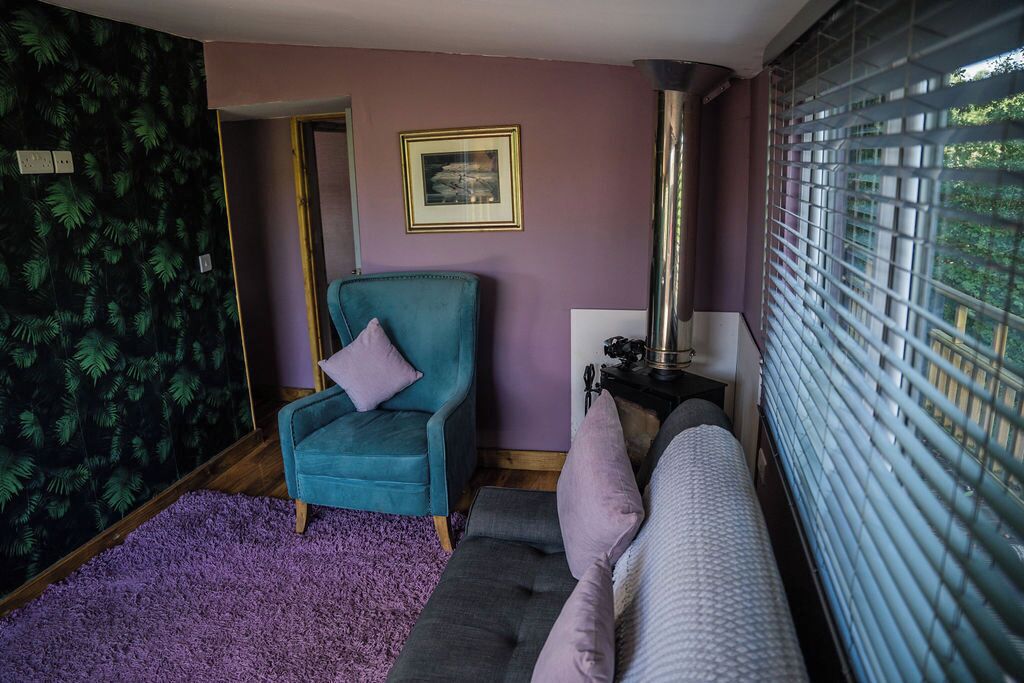 Una habitación con un sillón verde azulado, un sofá morado, un cuadro en la pared y una ventana con persianas.