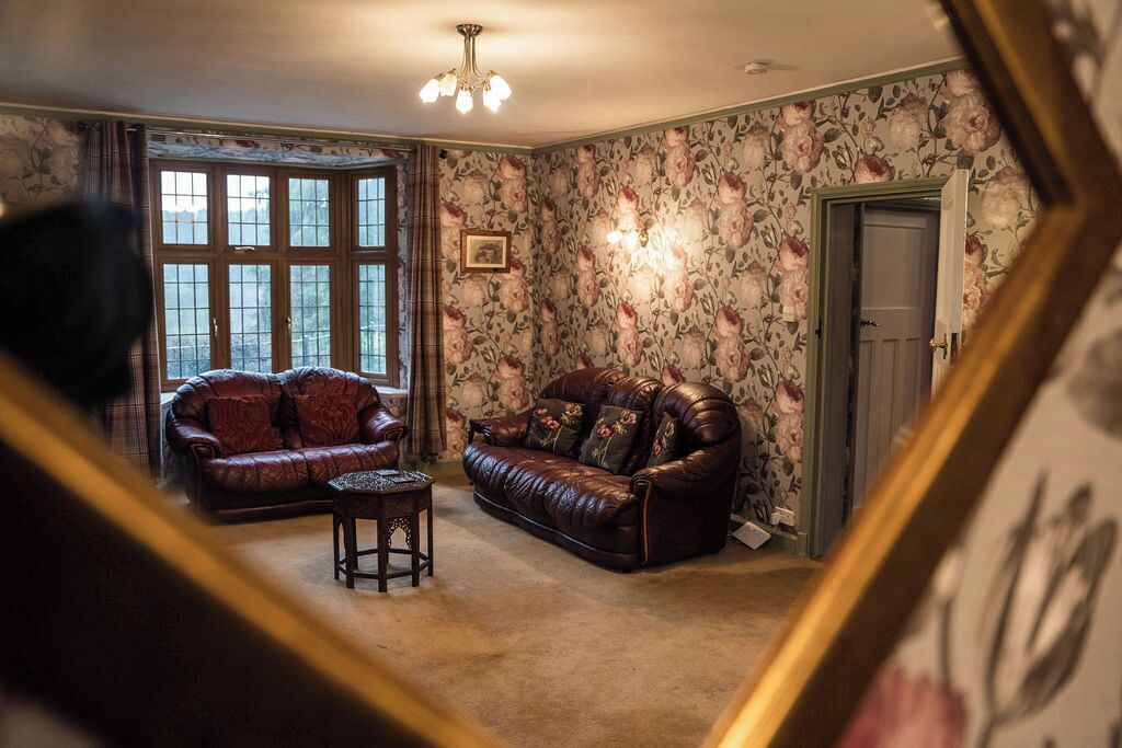 Een kamer met twee leren banken, een kleine tafel, bloemenbehang, een kroonluchter en een groot raam.