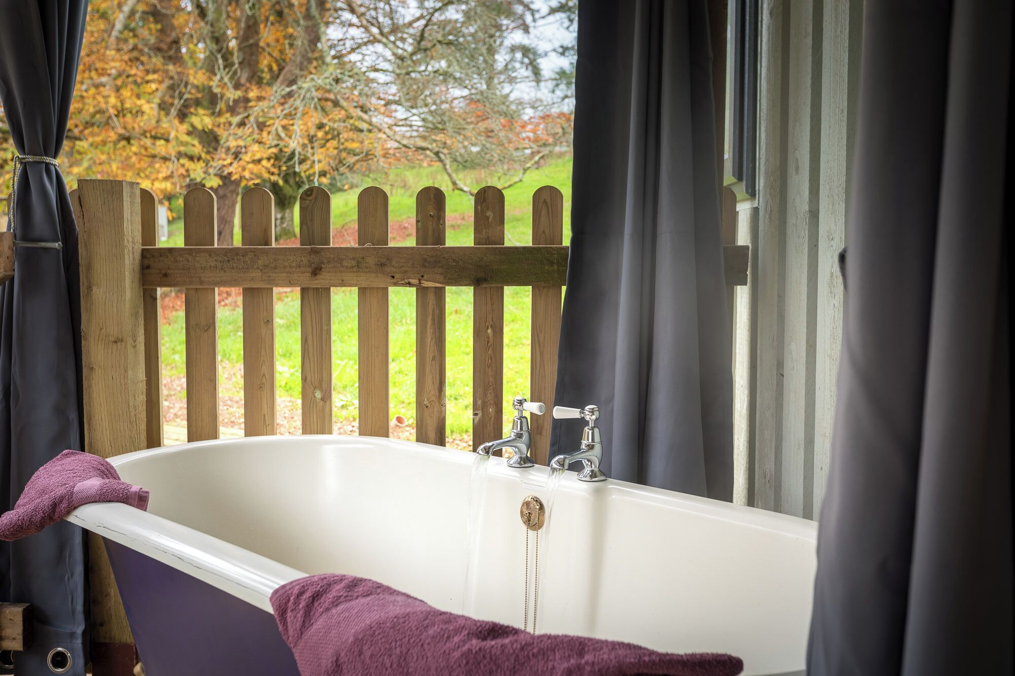 A bathroom with a white bathtub, a wooden fence outside the window, and a purple towel on the edge.