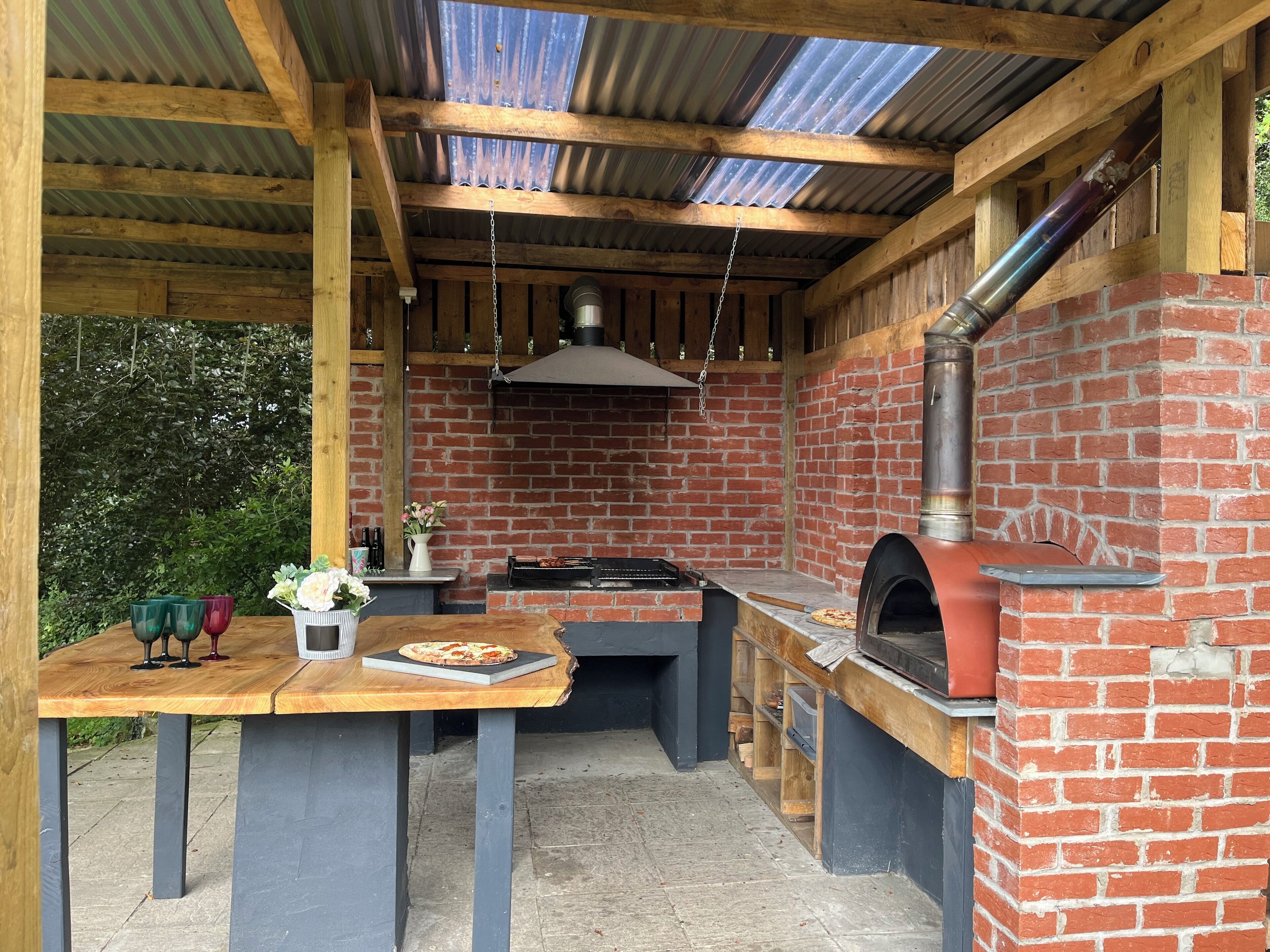 Une cuisine extérieure dotée d’un four en briques, d’un espace dédié à la préparation de pizzas et d’une table en bois où sont disposées des boissons et des fleurs.