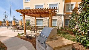 En udendørs patio med grill, siddepladser og en swimmingpool i baggrunden.