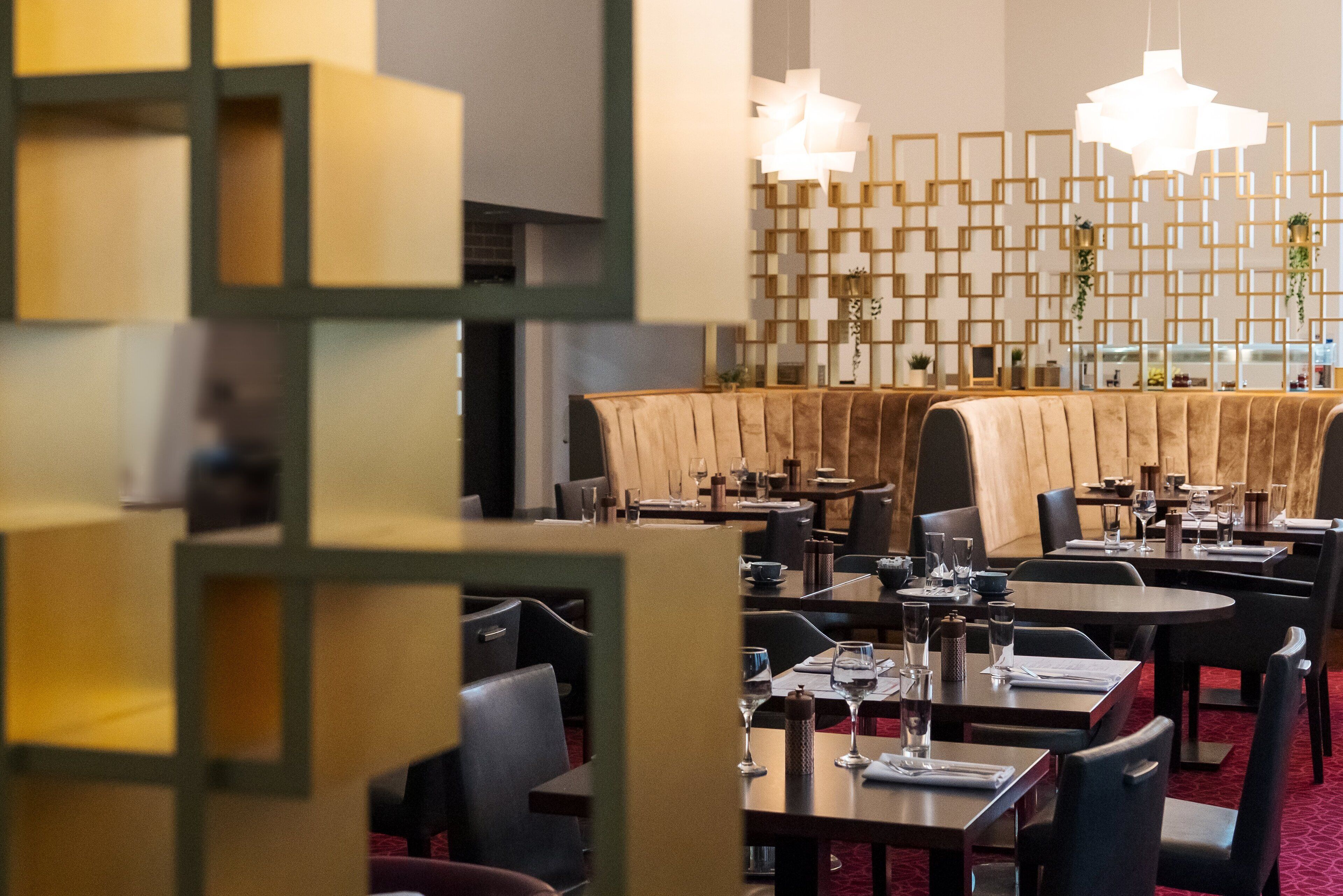 Een modern restaurant met meerdere eettafels en stoelen, een decoratieve muur met geometrische patronen en hangende pendelverlichting.