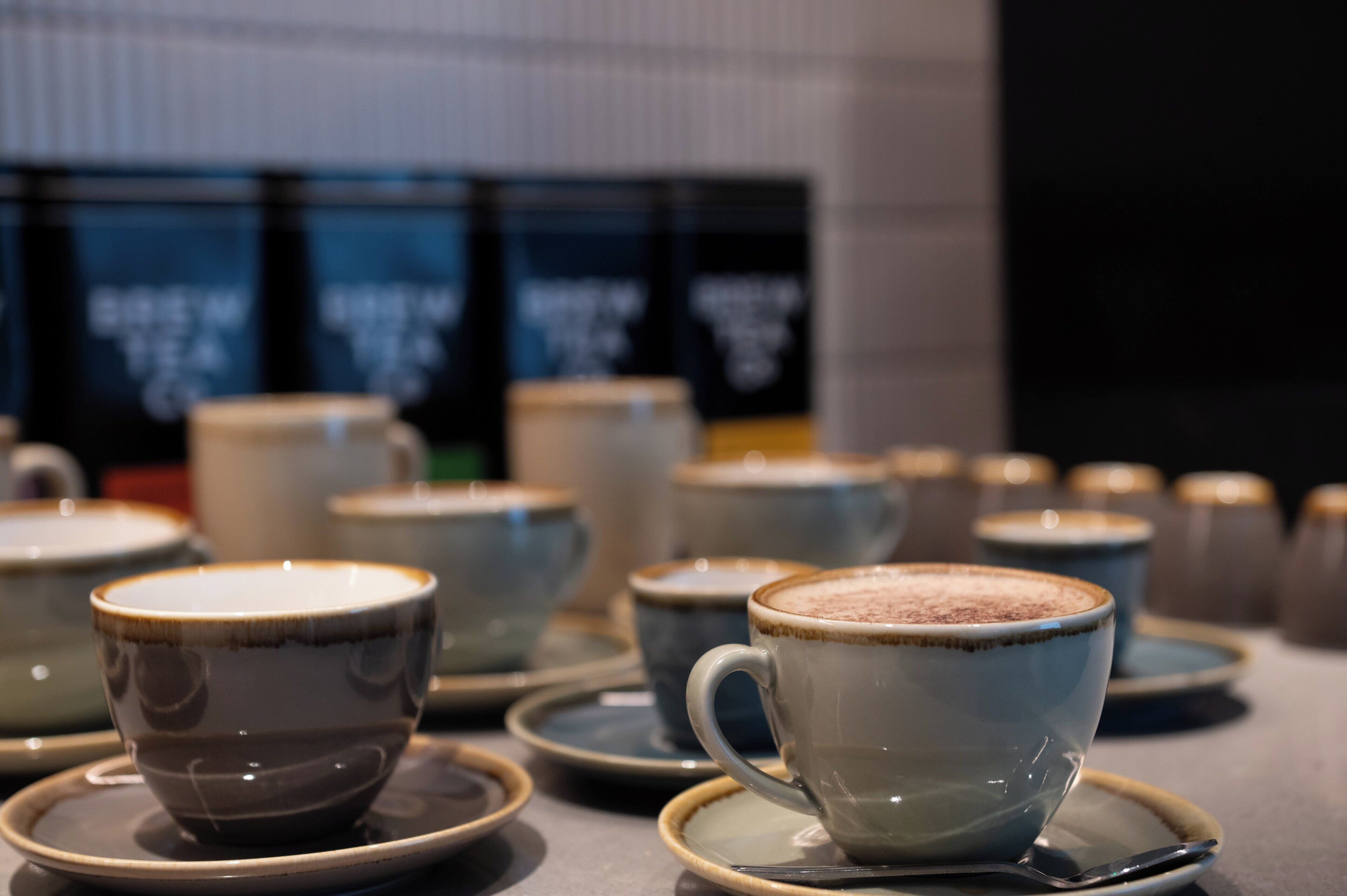 Una fila de tazas de cerámica con sus platillos, algunas con una bebida espumosa, dispuestas sobre una mesa.