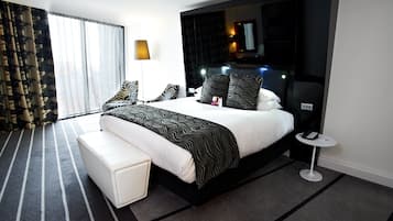 A modern hotel room with a large bed, a bedside table, a small side table, a large window with curtains, and a headboard with built-in lighting.