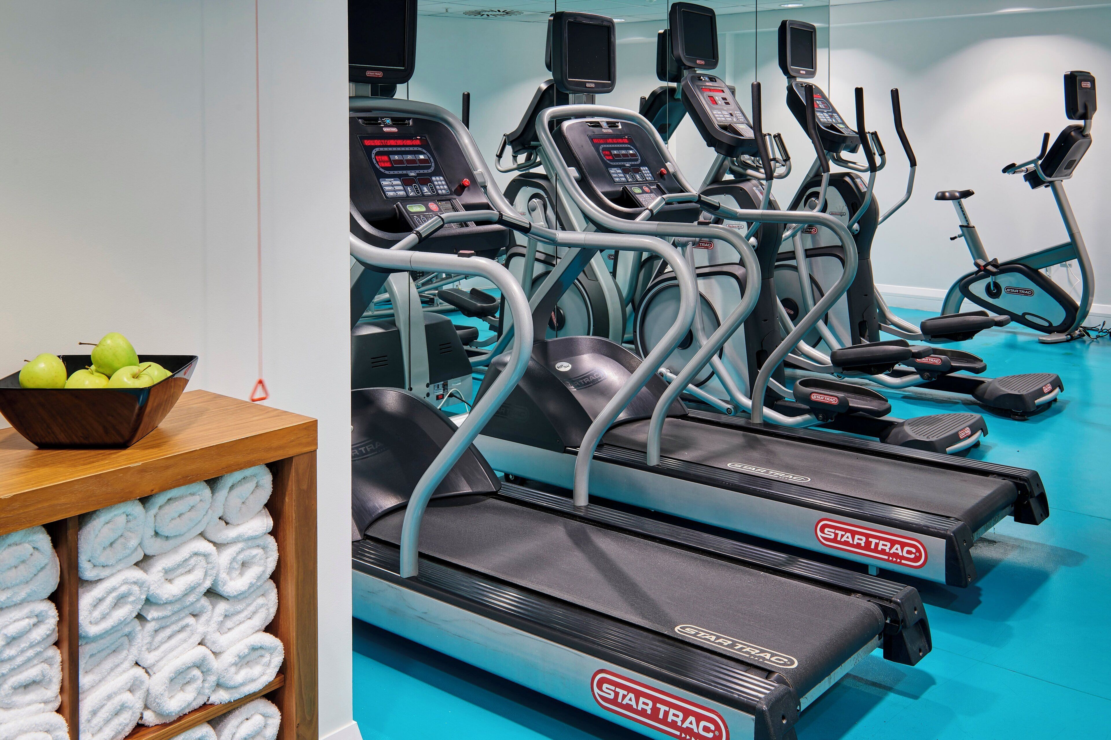Un gimnasio con varias cintas de correr, bicicletas estáticas y una mesa de madera con toallas y manzanas.