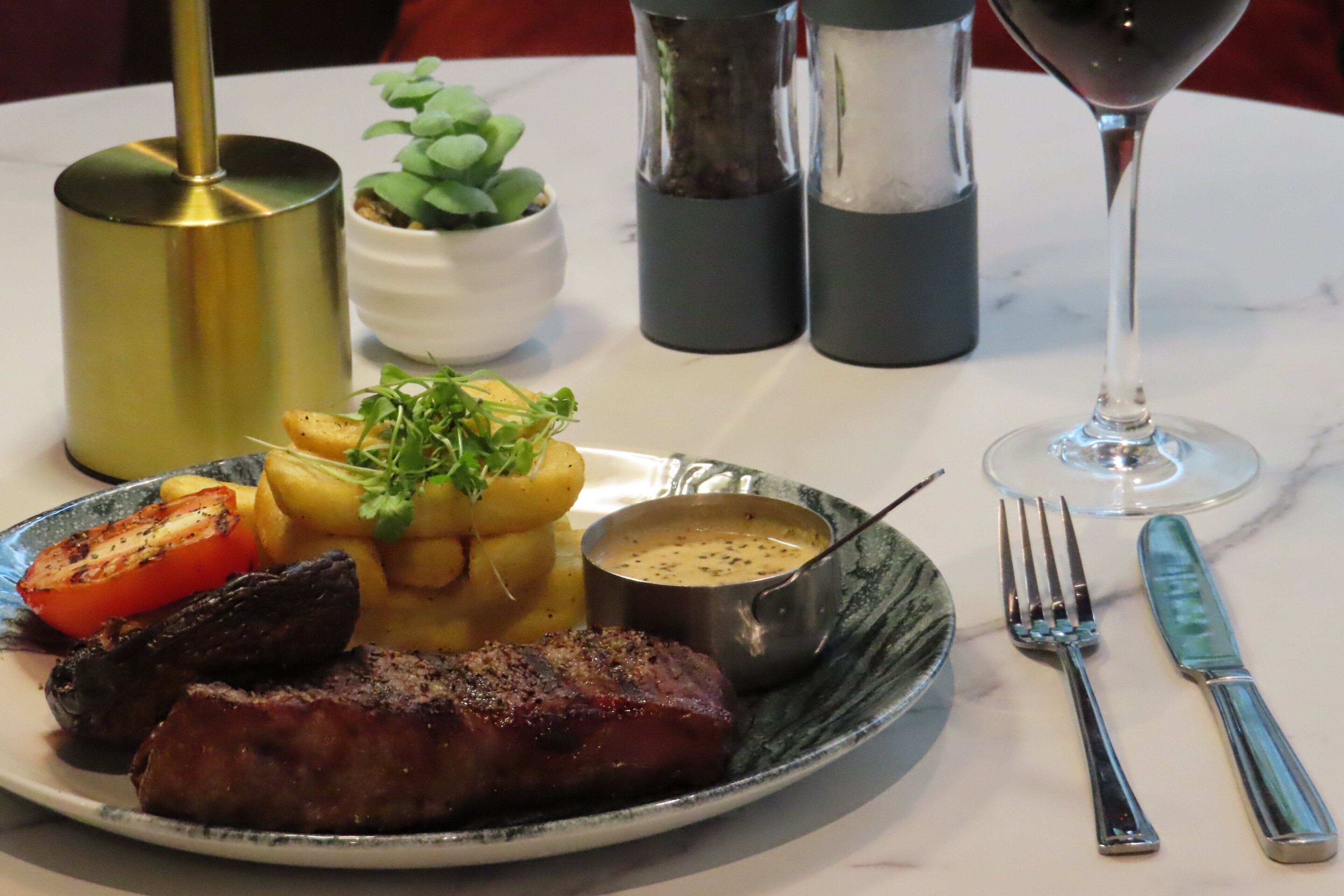 A plate of food with a steak, roasted potatoes, and a tomato, accompanied by a glass of wine and cutlery on a table.
