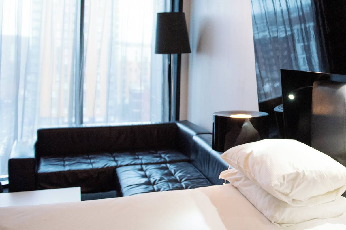 Een moderne hotelkamer met een zwarte leren bank, een wit bed en een groot raam met uitzicht over de stad.