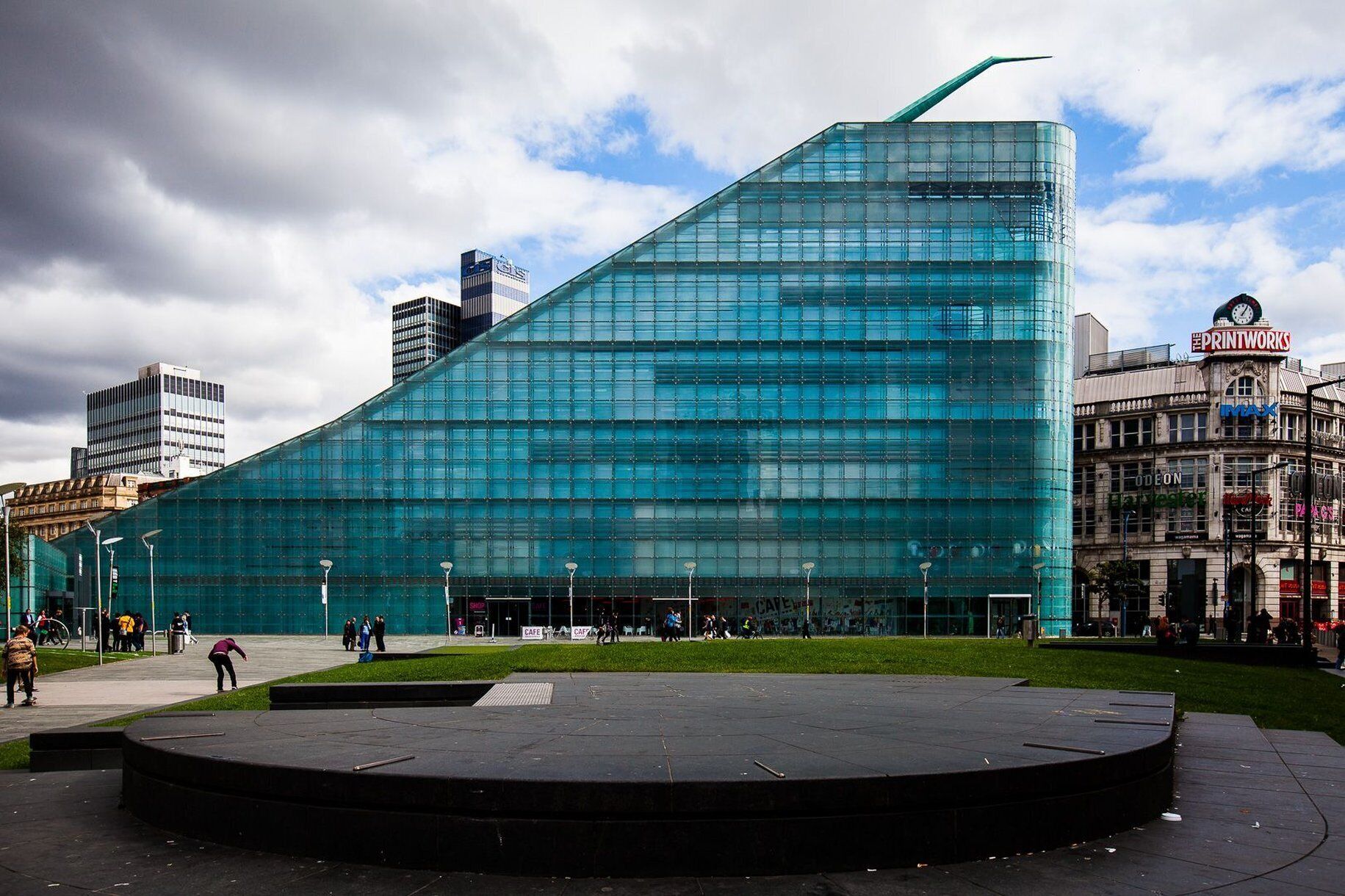 Un edificio moderno de vidrio con un diseño angular distintivo, rodeado por una plaza pública con un escenario circular.