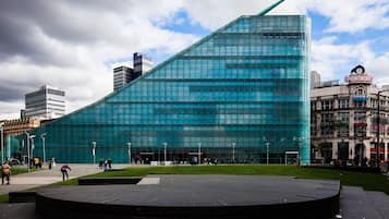 Un edificio moderno de vidrio con un diseño angular distintivo, rodeado por una plaza pública con un escenario circular.
