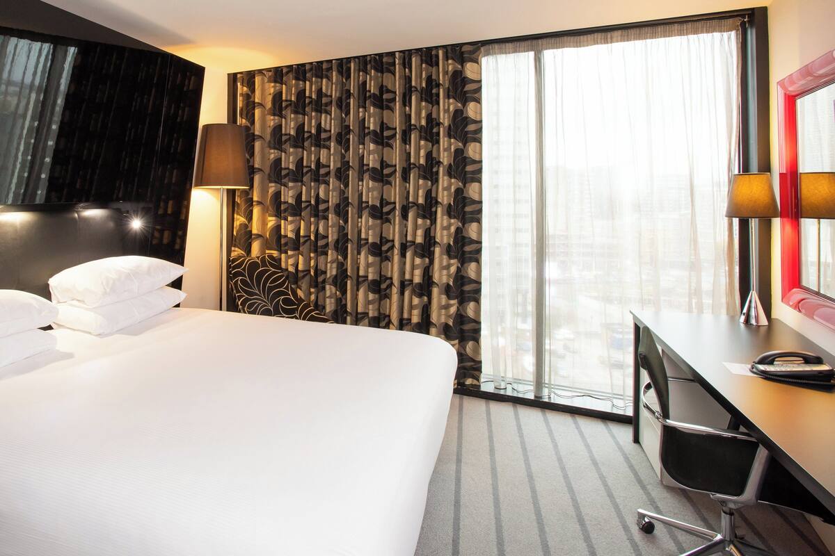 Een hotelkamer met een groot bed, een bureau met stoel, een lamp en uitzicht op de stad door dunne gordijnen.