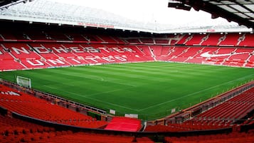 A stadium with red seating and a green field, featuring the Manchester United logo.