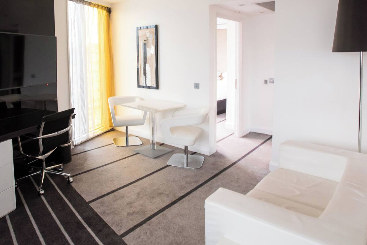Een moderne hotelkamer met een flatscreen televisie, een bureau met stoel, een witte bank en een eettafel met stoelen.