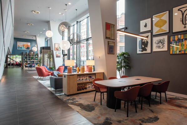 Un moderno vestíbulo de hotel con un mostrador de recepción de madera, sillas rojas y negras, una mesa redonda grande y una mezcla de arte contemporáneo y clásico en las paredes.