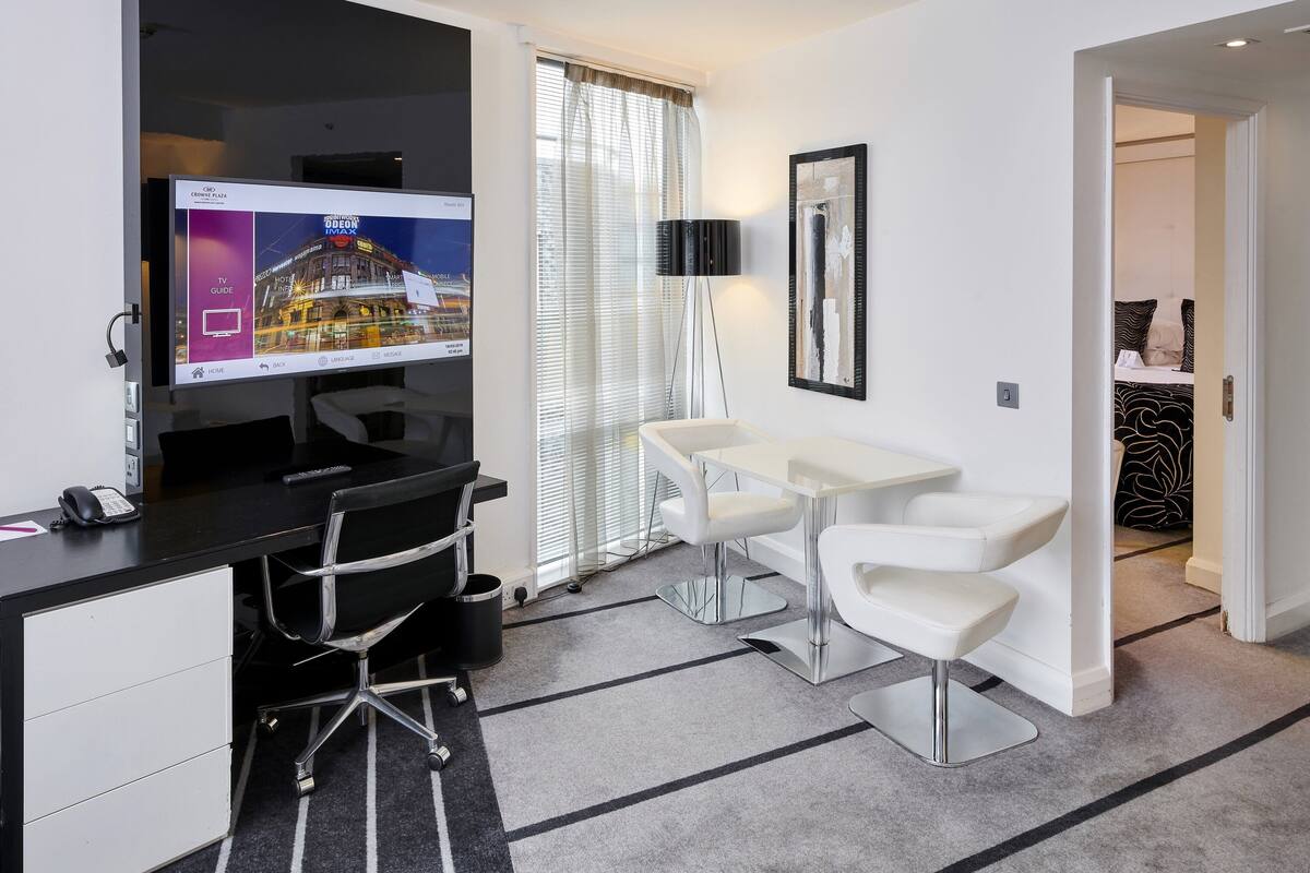 Een moderne hotelkamer met een bureau, een flatscreen televisie, een zithoek met een witte tafel en stoelen, en een groot raam met zonwering.