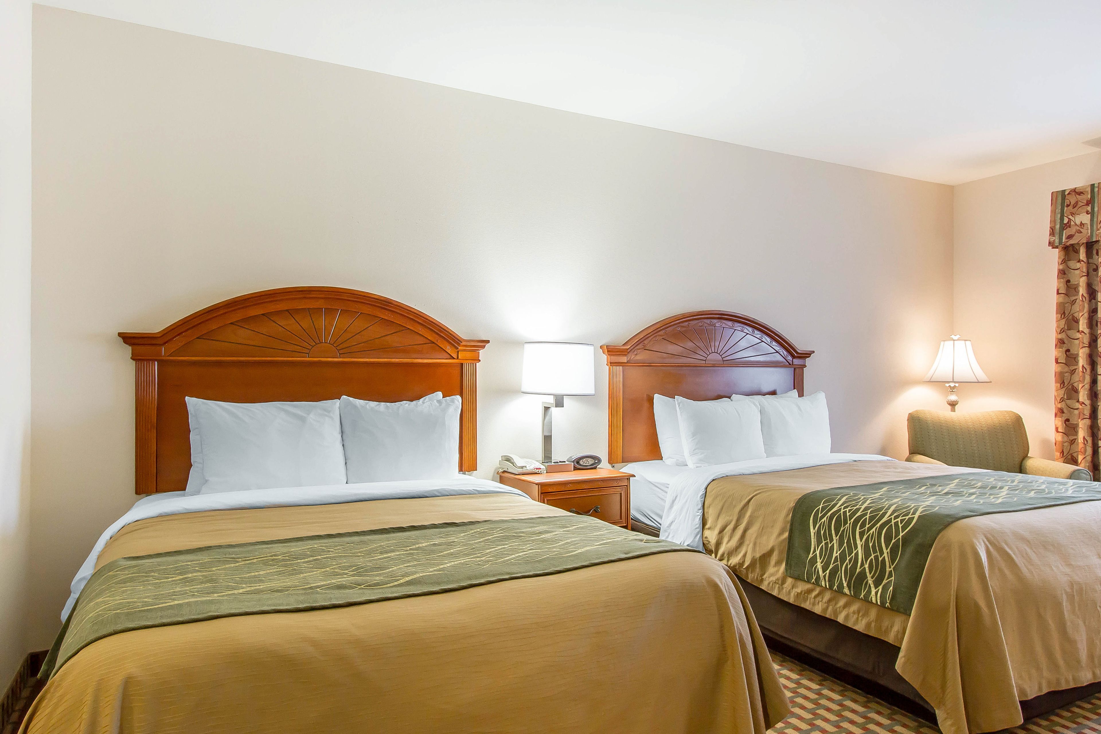 A hotel room with two beds, each with wooden headboards, white bedding, and a green throw blanket. There are bedside tables with lamps and a small clock.