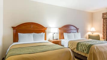 A hotel room with two beds, each with wooden headboards, white bedding, and a green throw blanket. There are bedside tables with lamps and a small clock.