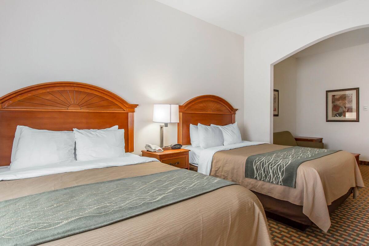 Camera d'albergo con due letti, entrambi con testiera in legno, biancheria da letto bianca e una coperta decorata. Si vede un comodino con una lampada e un telefono, oltre a un tavolino con una sedia.