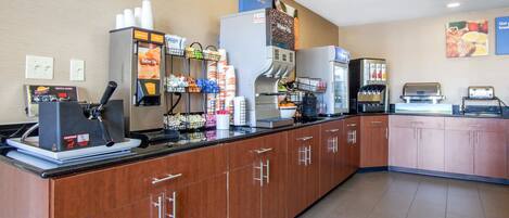 Un área de desayuno autoservicio con máquinas de café, un refrigerador y diversos alimentos.