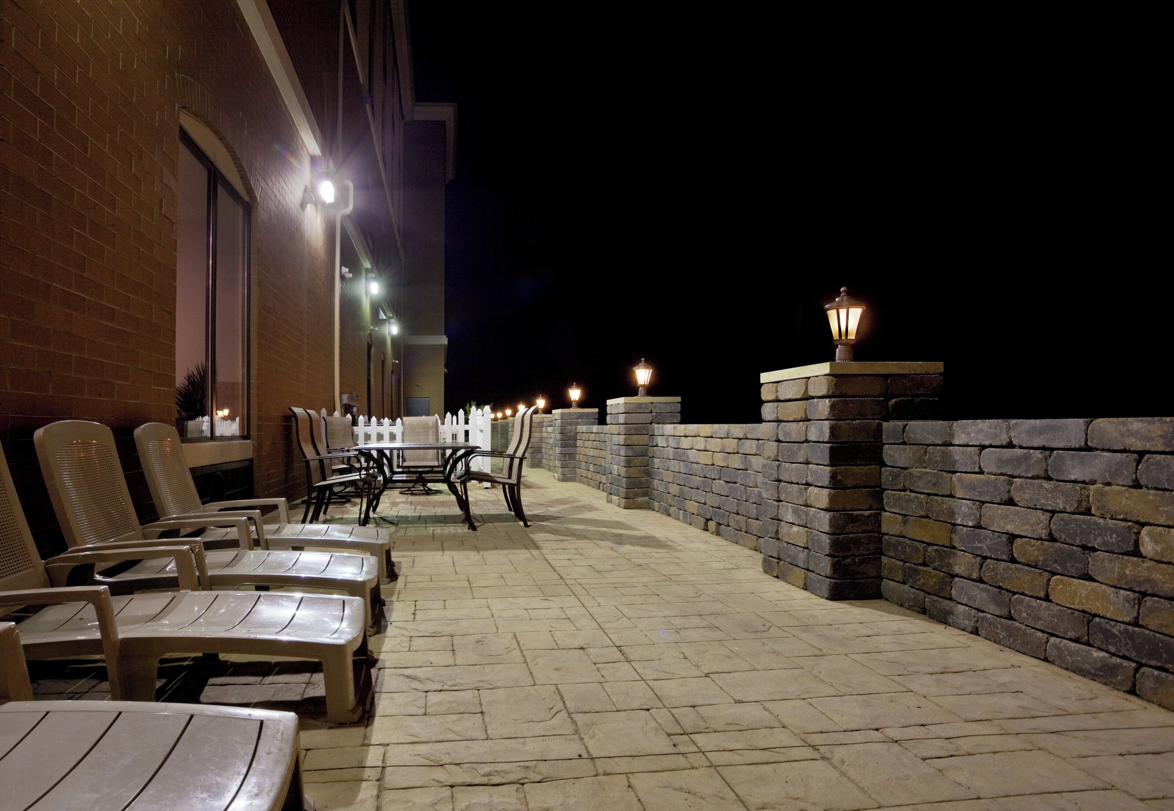Un espacio al aire libre con mesas y sillas, un muro de ladrillo e iluminación decorativa.