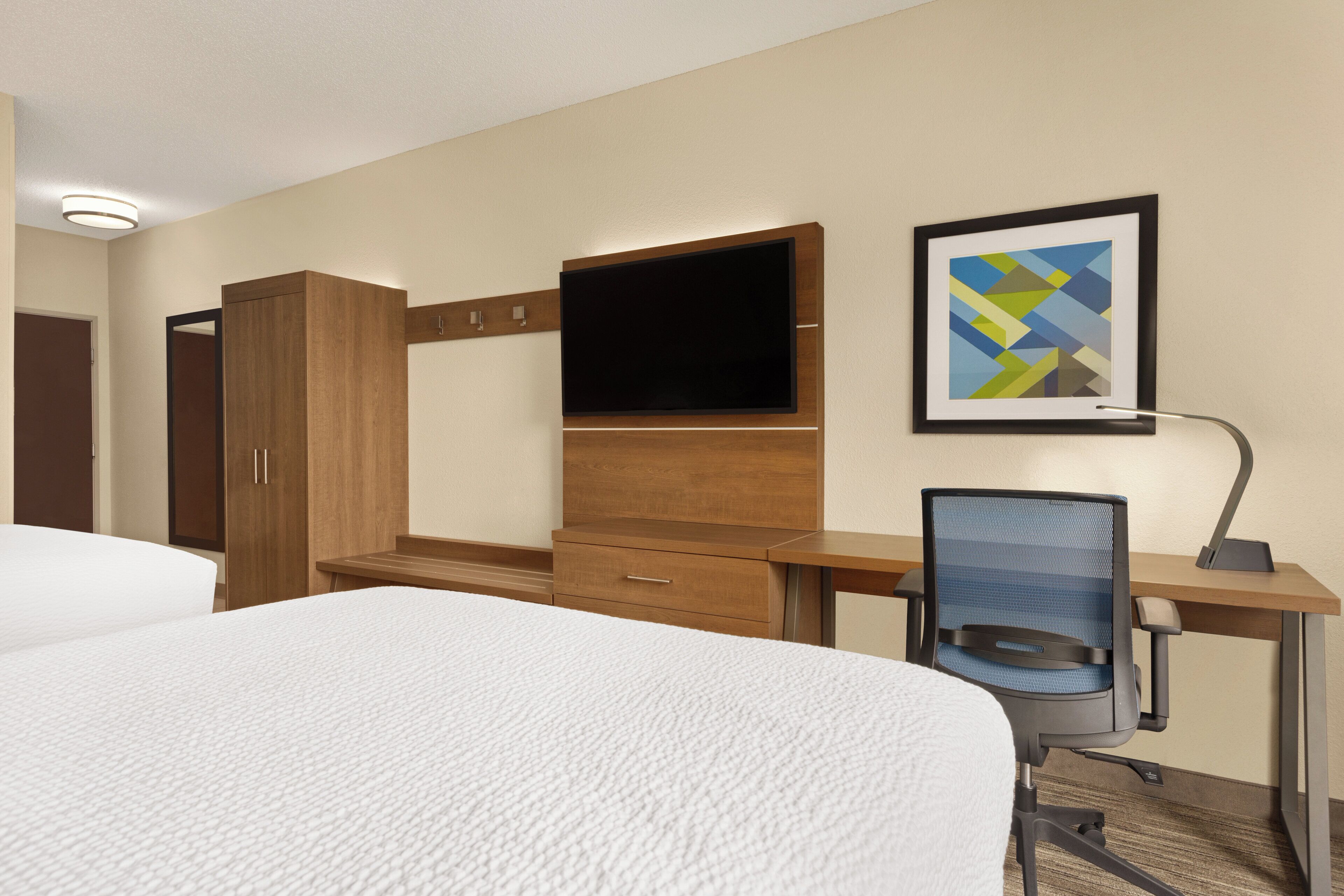 A hotel room with two beds, a desk with a chair, a flat-screen TV, a wooden wardrobe, and a framed picture on the wall.