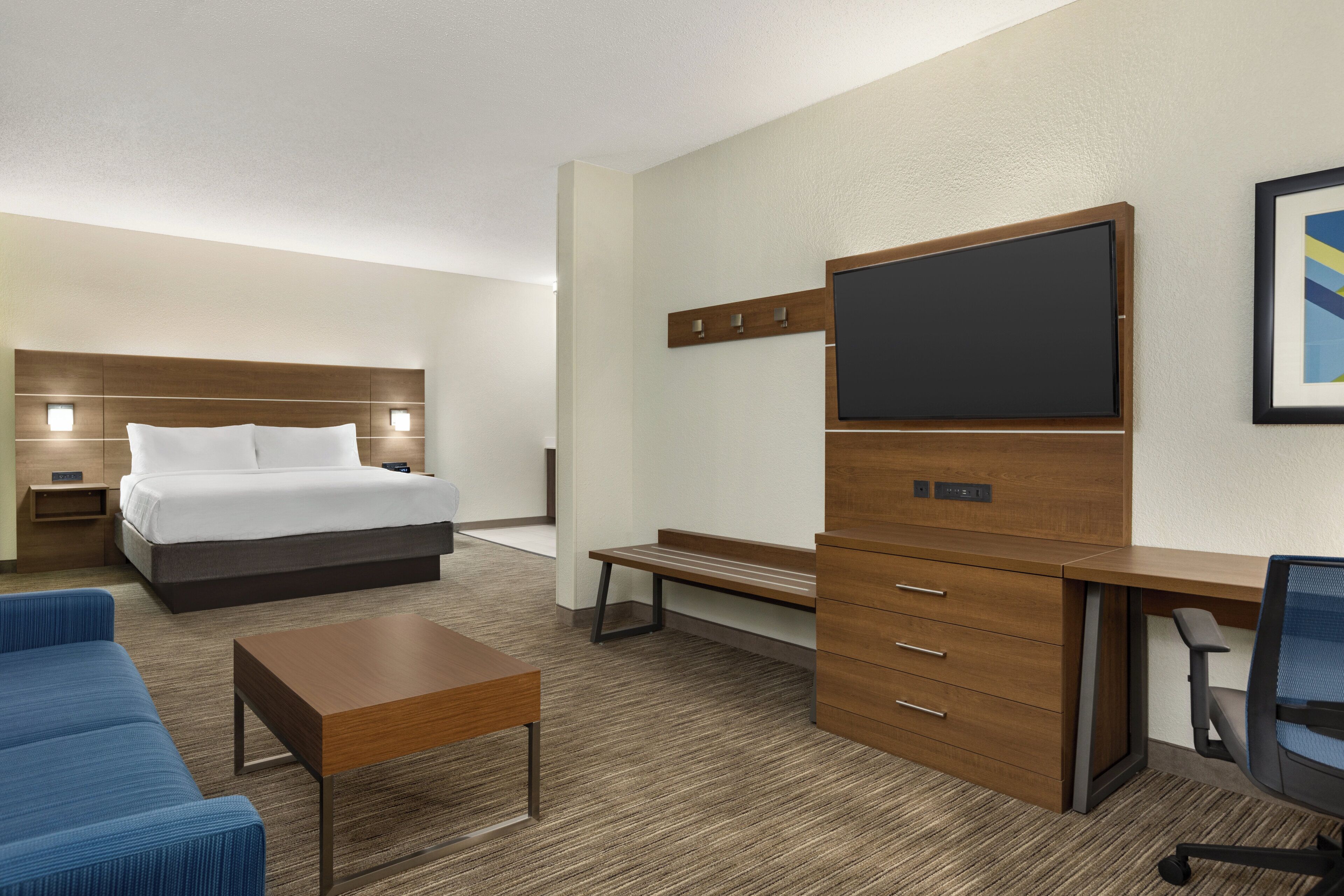 A hotel room with a bed, a desk with drawers, a chair, a small table, and a TV mounted on the wall.