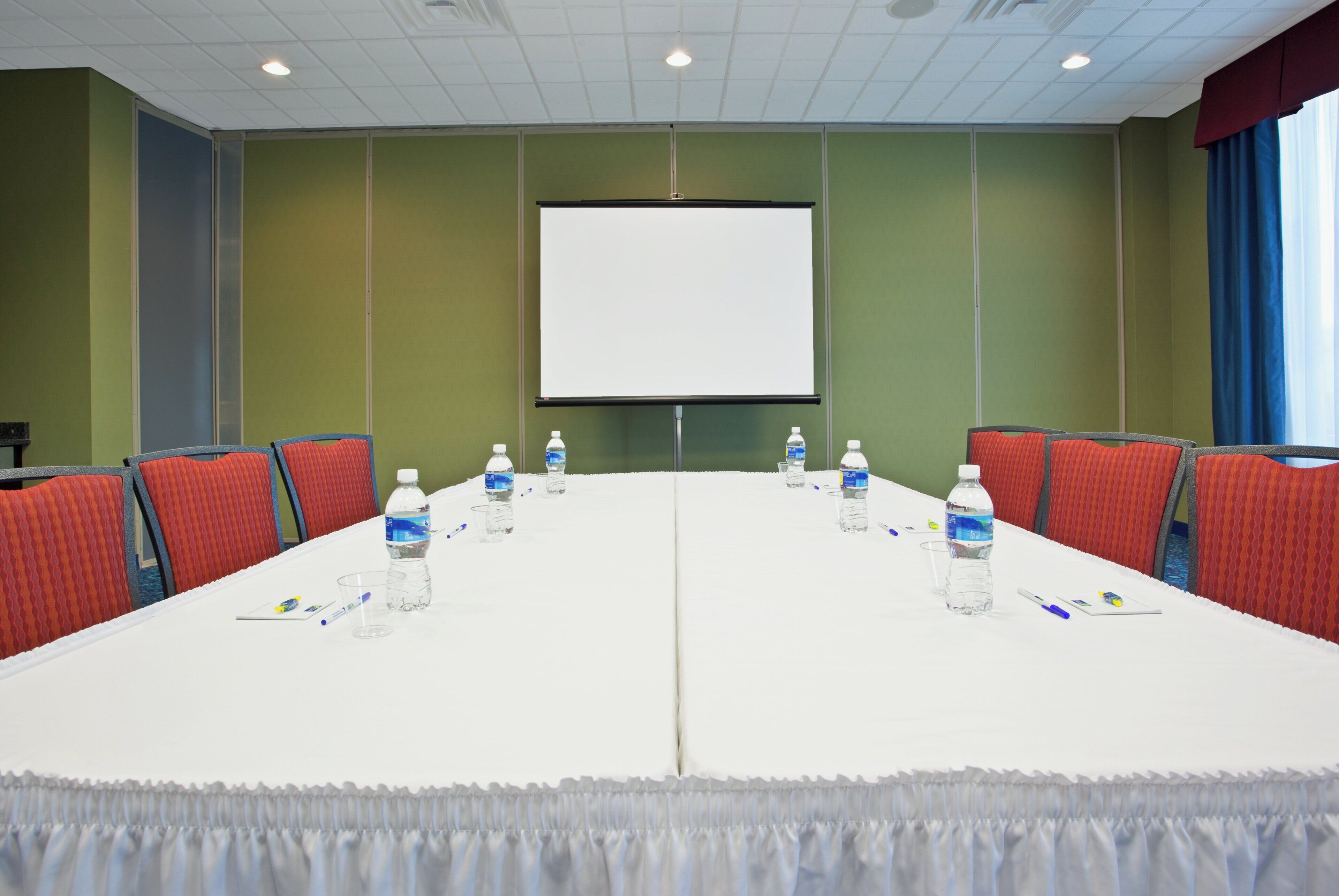 A conference room with a long table, chairs, water bottles, and a projector screen.