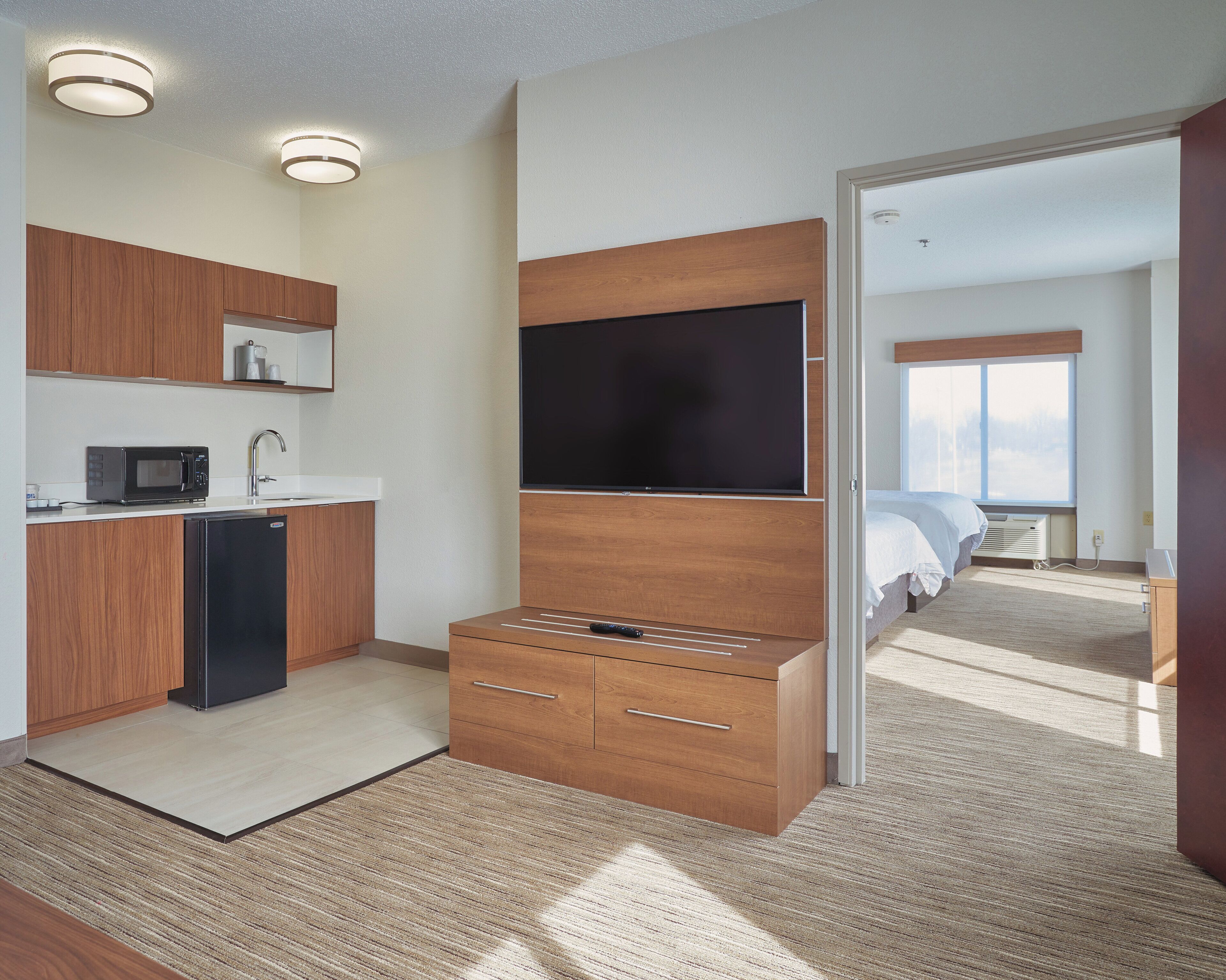 Une chambre d’hôtel moderne dotée d’une kitchenette, d’un téléviseur à écran plat, d’une commode en bois et d’une grande fenêtre offrant une vue panoramique.