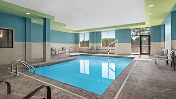 A swimming pool with a tiled deck, chairs, and a view of trees through large windows.