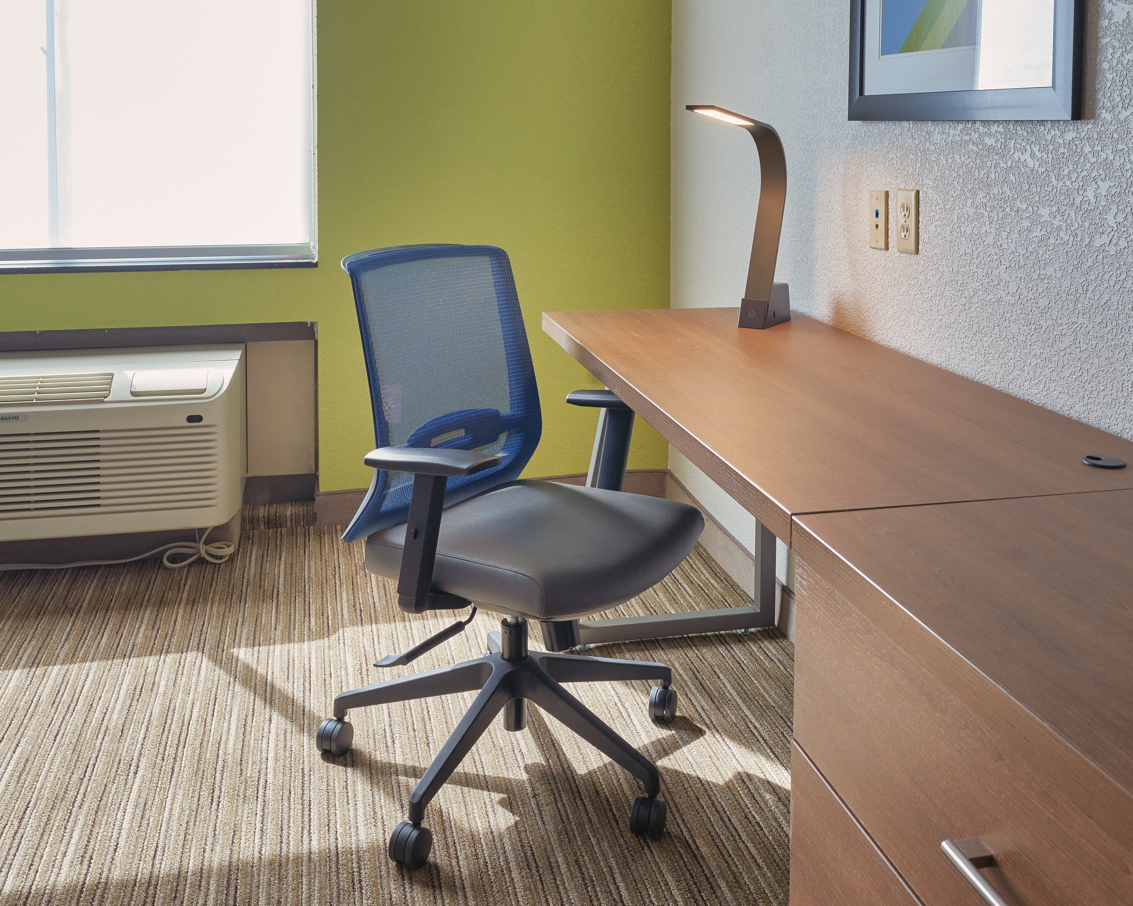 A room with a desk, chair, standing lamp, air conditioning unit, and a window.
