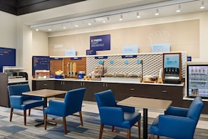 A breakfast area with a counter displaying various food options, a coffee machine, and seating with blue chairs and tables. - Holiday Inn Express & Suites Akron Regional Airport Area by IHG (Akron)