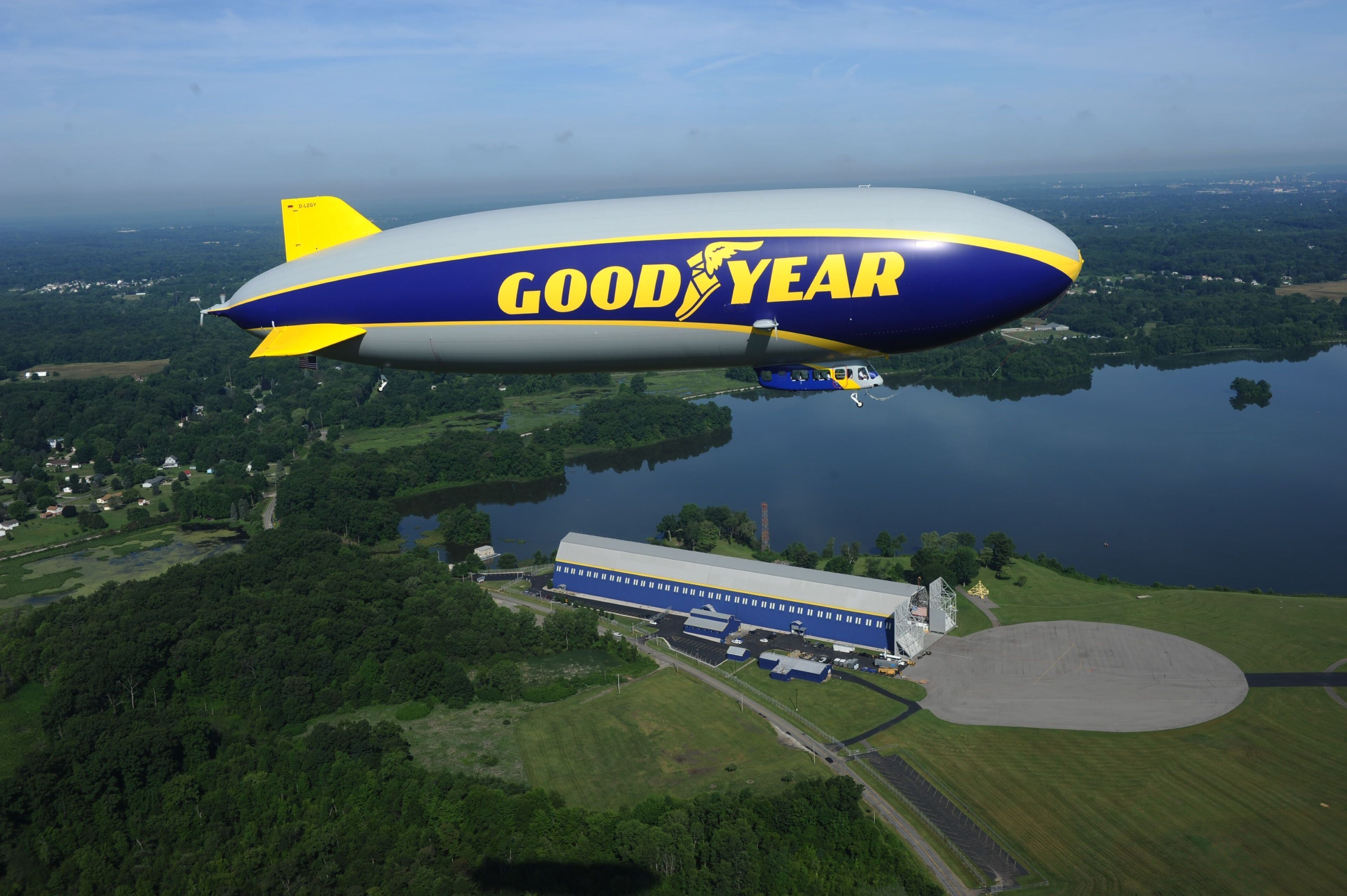 A Goodyear blimp flying over a rural area with a large building below.