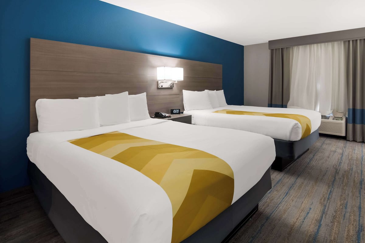 Ein Hotelzimmer mit zwei Betten, einem Nachttisch, einer Lampe, einer Klimaanlage und einem Fenster mit Vorhängen.