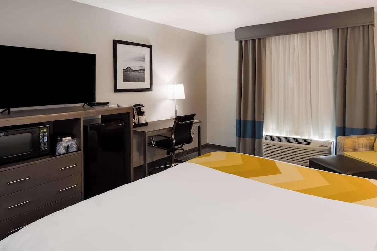 Ein Hotelzimmer mit einem Bett, einem Schreibtisch, einem Fernseher, einer Mikrowelle, einem Kühlschrank, einem Sessel, einer Lampe und einem Fenster mit Vorhängen.