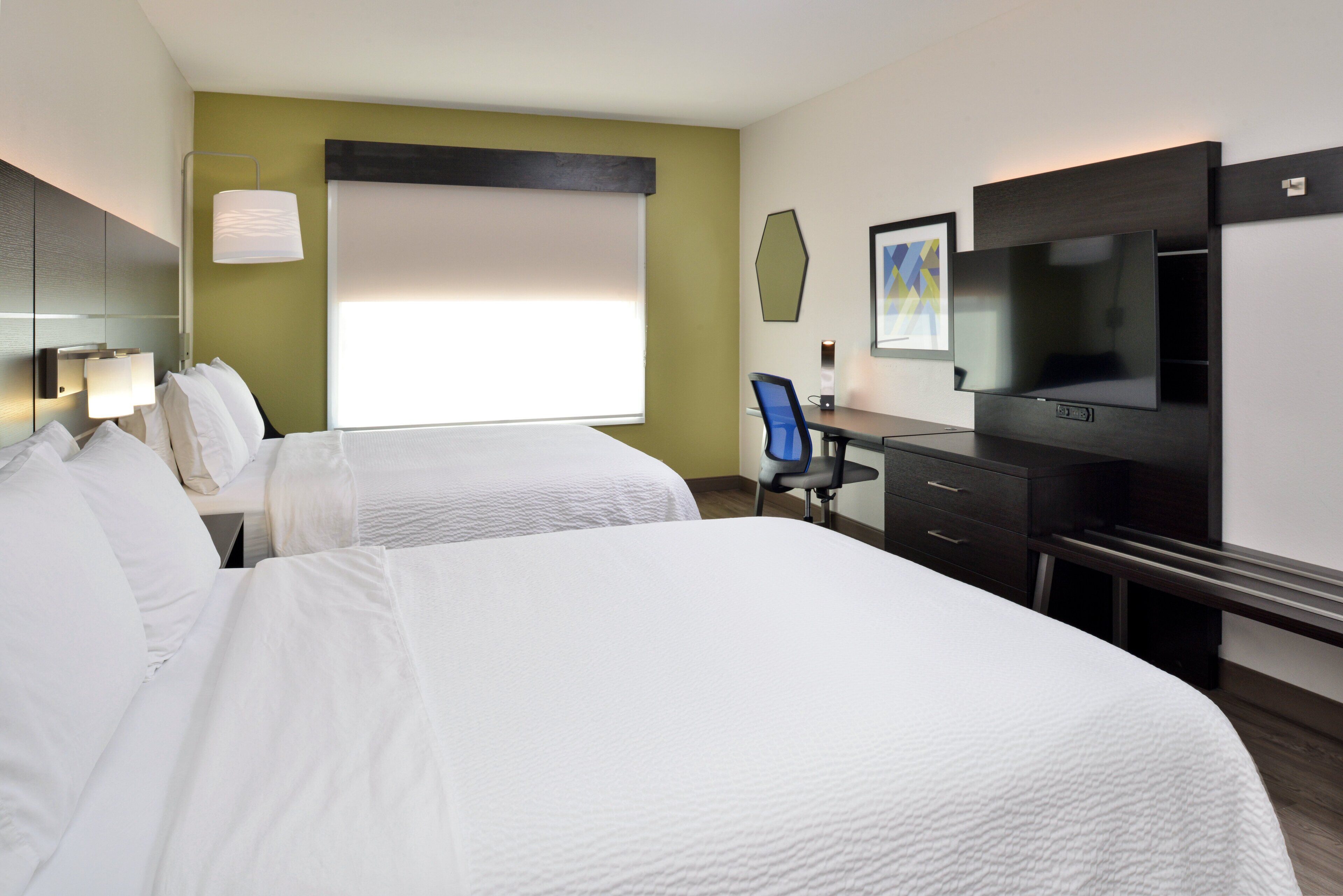 A hotel room with two beds, a desk, a chair, a TV, and a window with blinds.