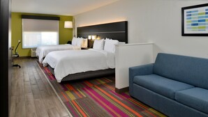 A hotel room with two beds, a blue sofa, a colorful striped carpet, and a large window with blinds.