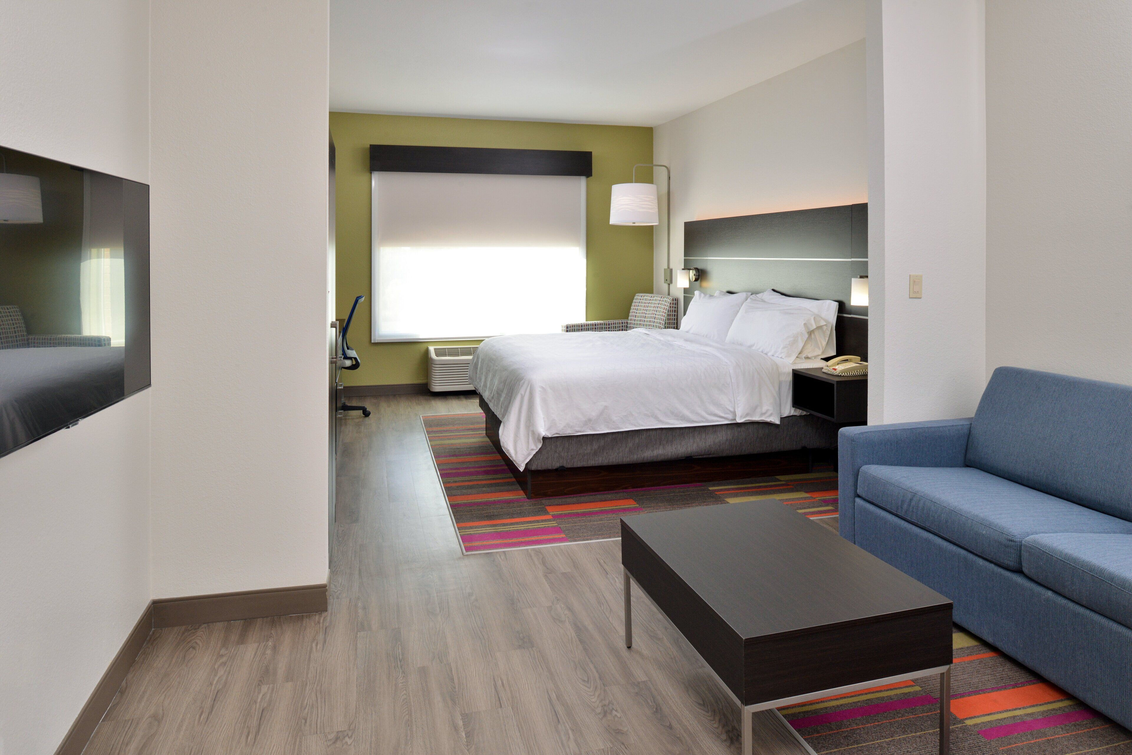 A hotel room with a bed, a blue sofa, a small table, a TV, and a window with blinds.
