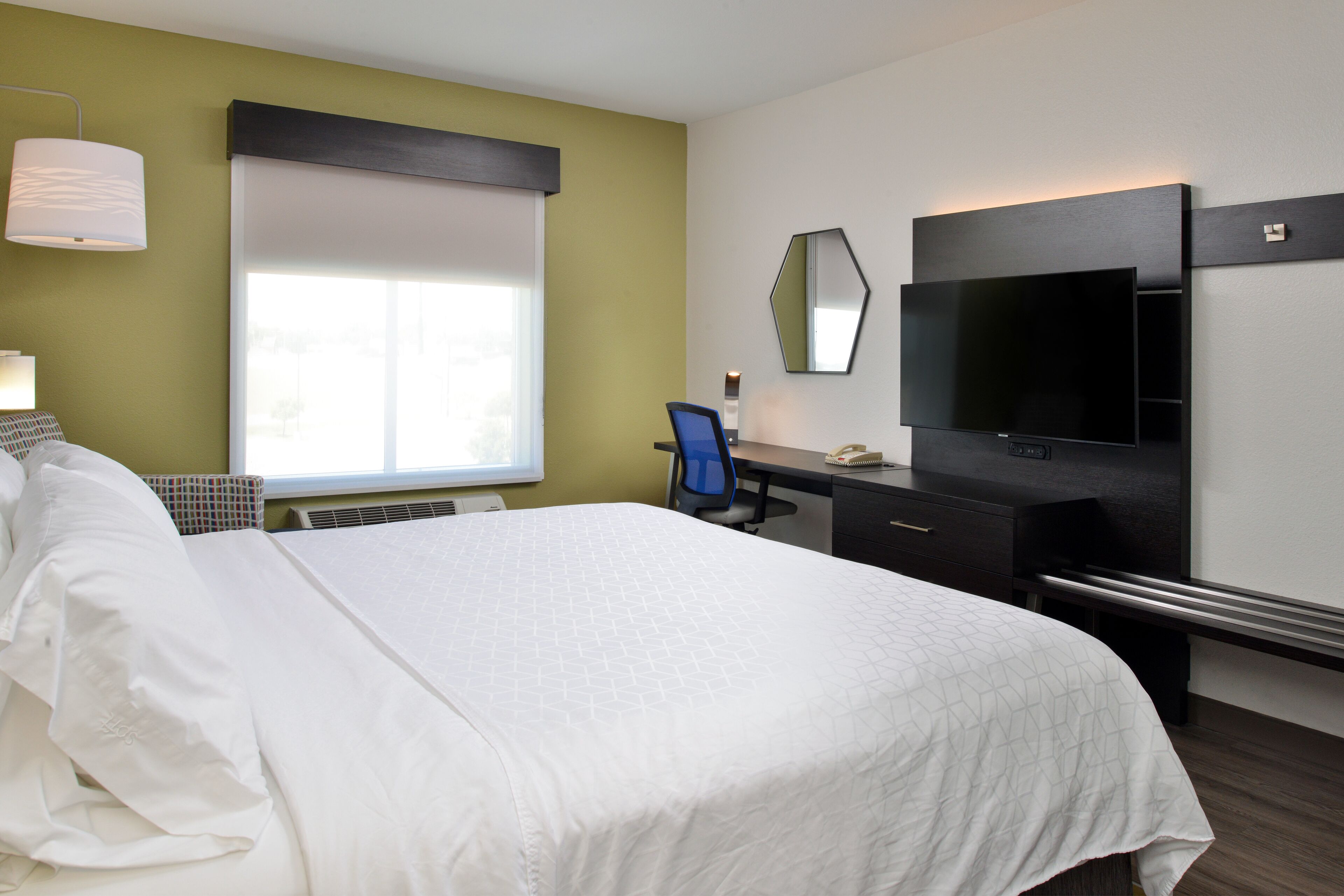 A hotel room with a large bed, a desk with a chair, a flat-screen TV, and a window with blinds.