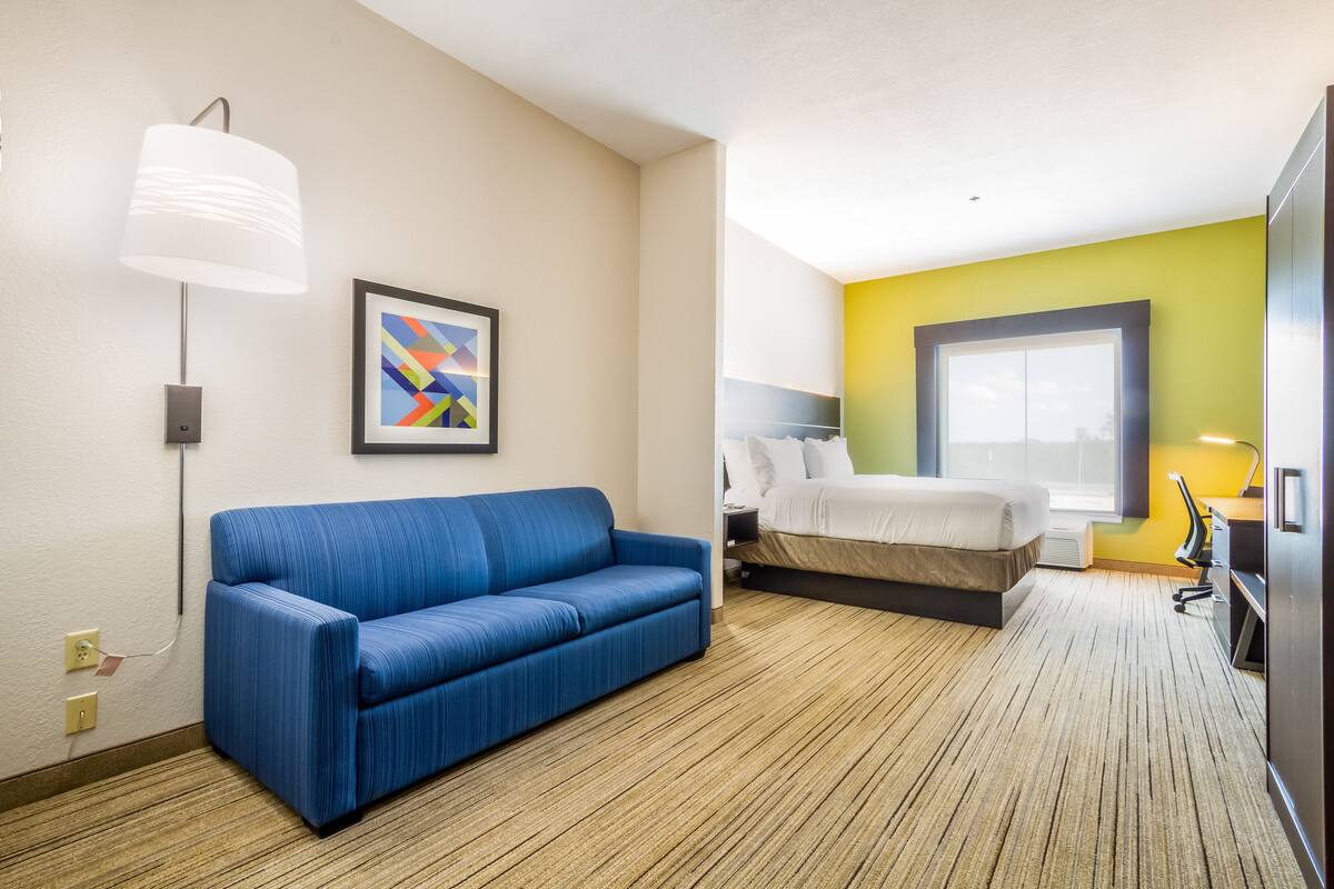 A hotel room with a blue sofa, a bed, a desk with a chair, a lamp, and a framed abstract painting.