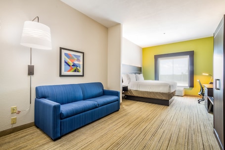 A hotel room with a blue sofa, a bed, a desk with a chair, a lamp, and a framed abstract painting.