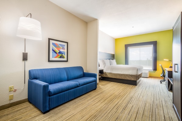 Une chambre d’hôtel avec un canapé bleu, un lit, un bureau avec une chaise, une lampe et un tableau abstrait encadré.