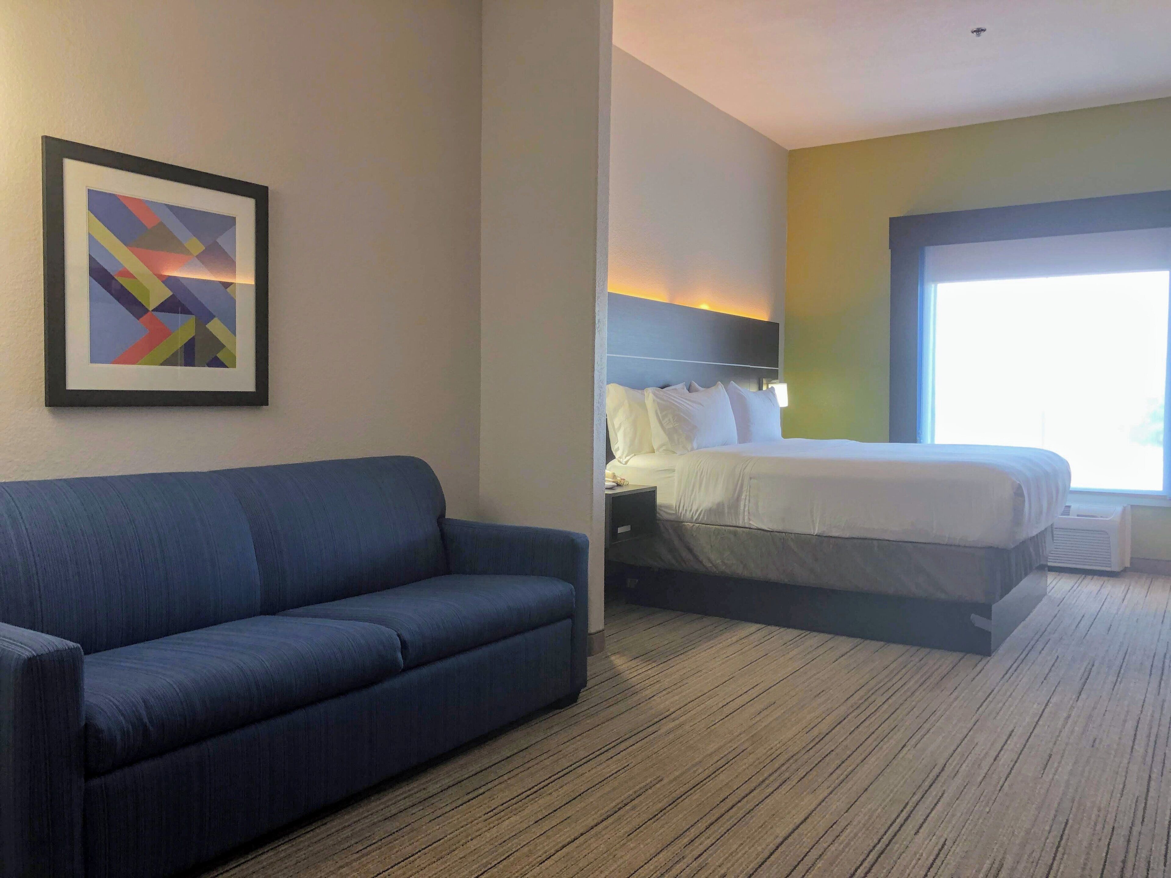 A hotel room with a blue sofa, a bed with white bedding, a nightstand, and a wall-mounted abstract painting.
