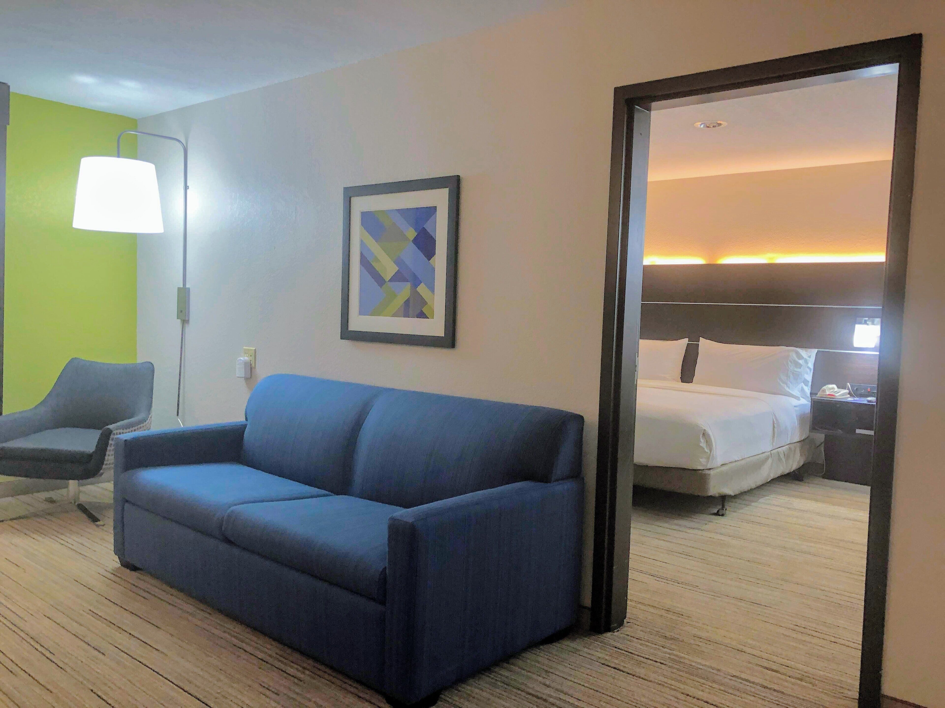 Une chambre d’hôtel avec un canapé bleu, une œuvre d’art abstraite encadrée, un lit avec du linge de maison blanc et une table de chevet avec une lampe.